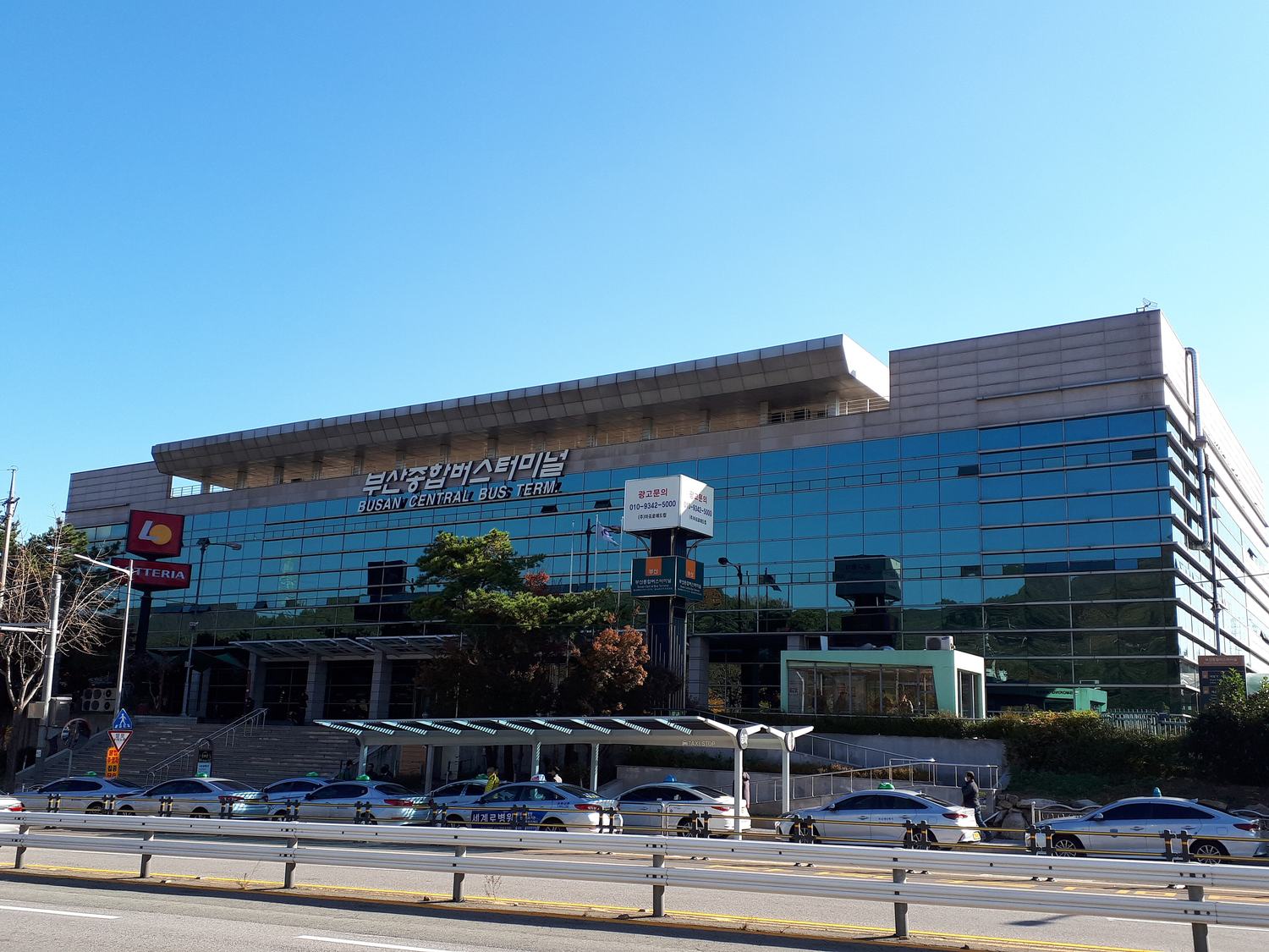 The glass-fronted Busan Central Bus Terminal building with 부산종합버스터미널 and BUSAN CENTRAL BUS TERMINAL on the facade, a row of taxis waiting at the stand in front, under a clear autumn sky.