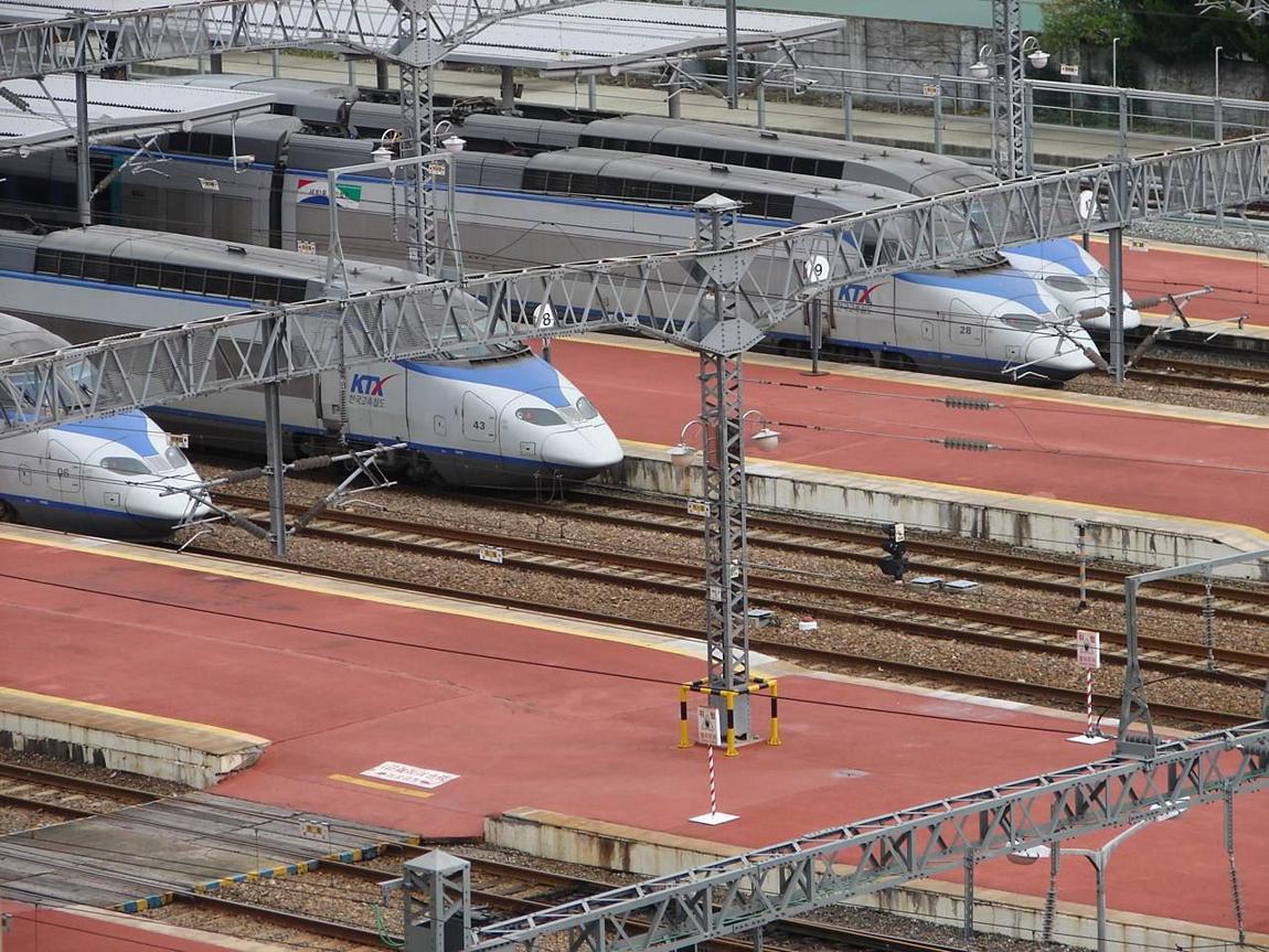 Several white-and-blue first-generation KTX trains parked side by side at the platforms of Busan Station, seen from above, with red-painted platforms and overhead line gantries.