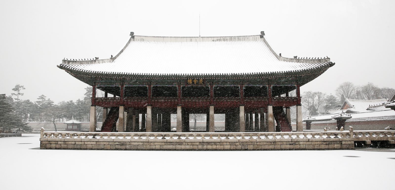 Gyeonghoeru, the two-storey banquet pavilion at Gyeongbokgung Palace in Seoul, with snow on its tiled roof and on the frozen pond in front, while snow is still falling.
