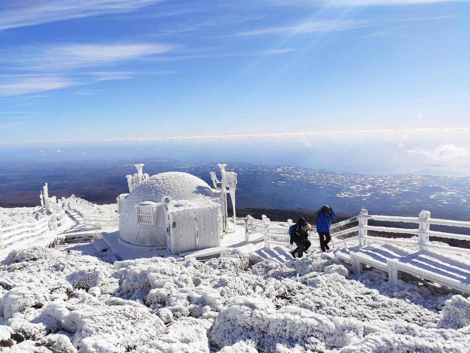 The summit area of Hallasan on Jeju coated in white rime ice, with a domed structure, frozen wooden railings and two hikers, looking north over the island to the sea.