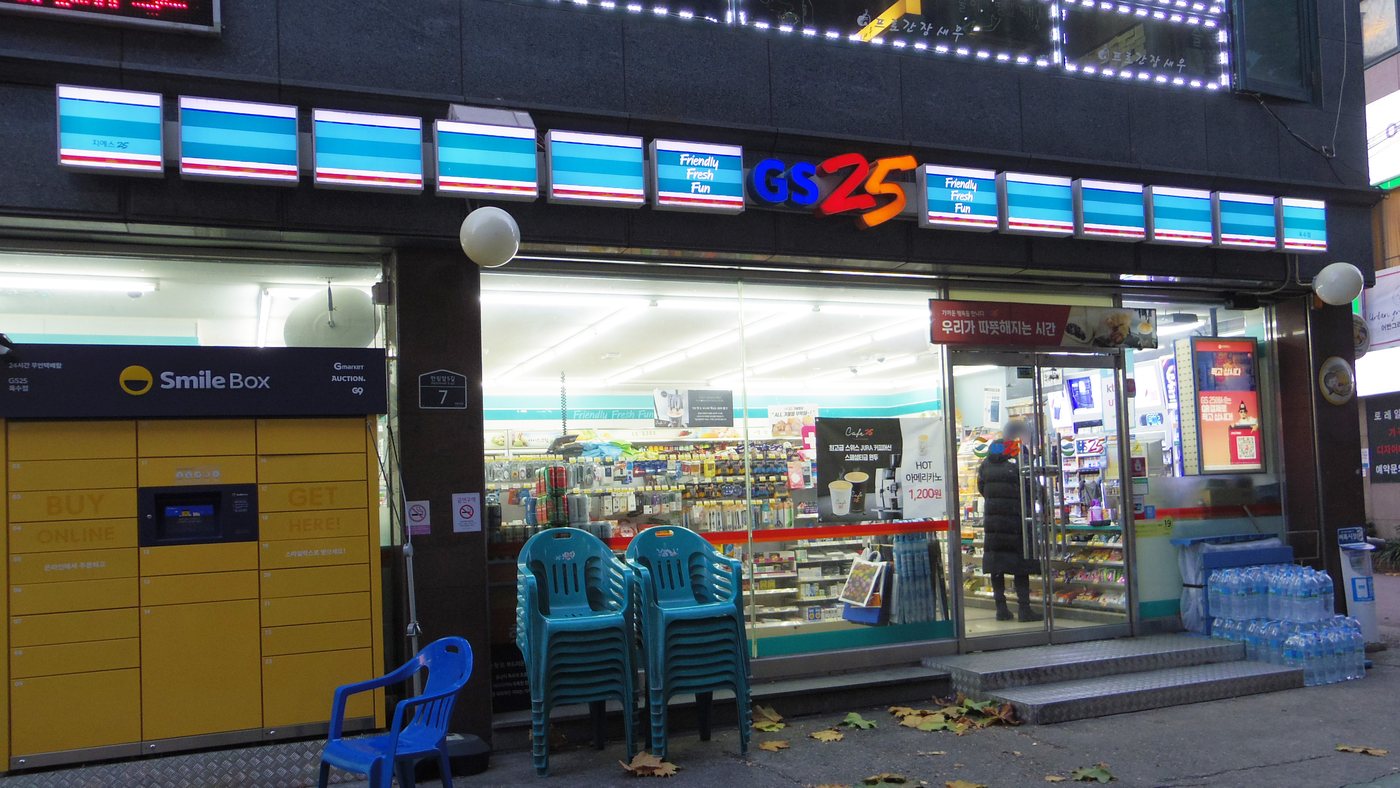 A GS25 convenience store at street level in Seoul in November, with a customer in a long padded coat at the door and stacked plastic chairs outside.