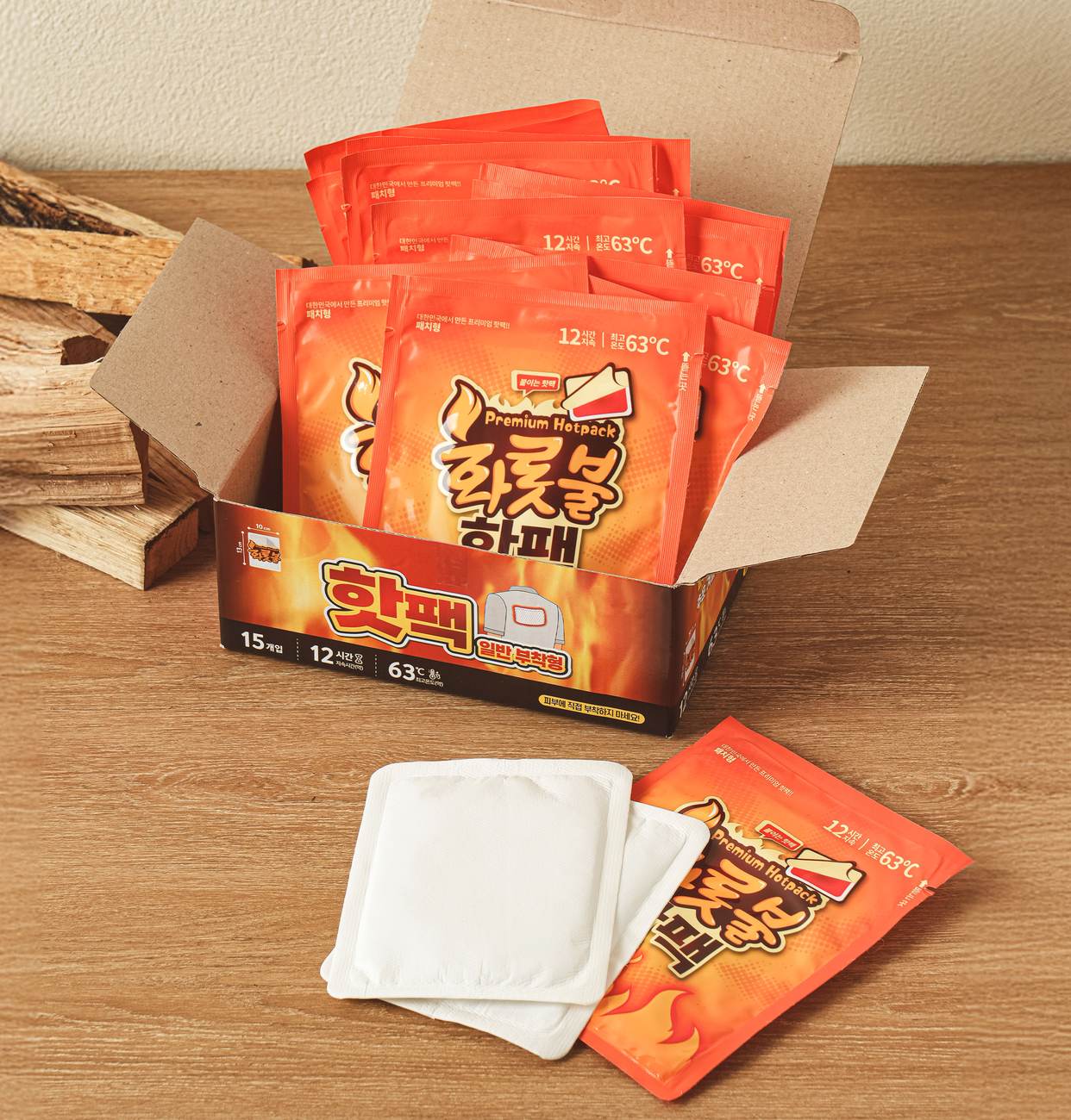 An open box of Daiso stick-on hot packs, 12 hours and 63°C, with orange 화롯불 핫팩 pouches standing inside and two white adhesive pads in front.