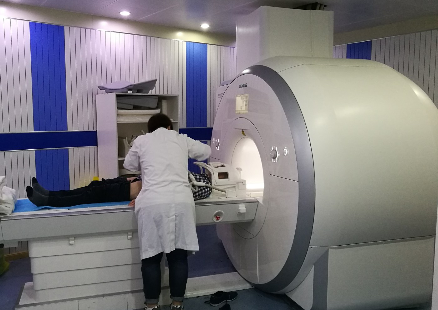 A radiographer in a white coat positions a patient's head in the head coil of a Siemens MRI scanner; the patient lies on the scanner table in a room with blue and white striped walls.