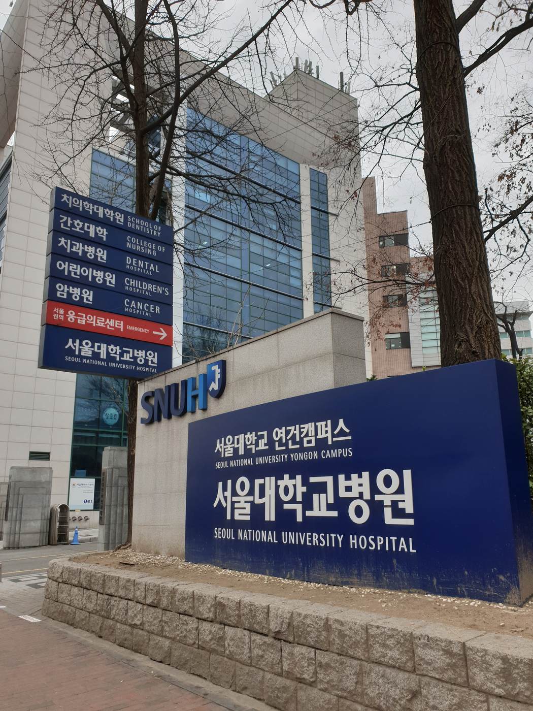 The front of Seoul National University Hospital: a blue stone-mounted sign reading 서울대학교병원 Seoul National University Hospital with the SNUH logo, and a directory post listing the dental hospital, children's hospital, cancer hospital and a red regional emergency centre sign.