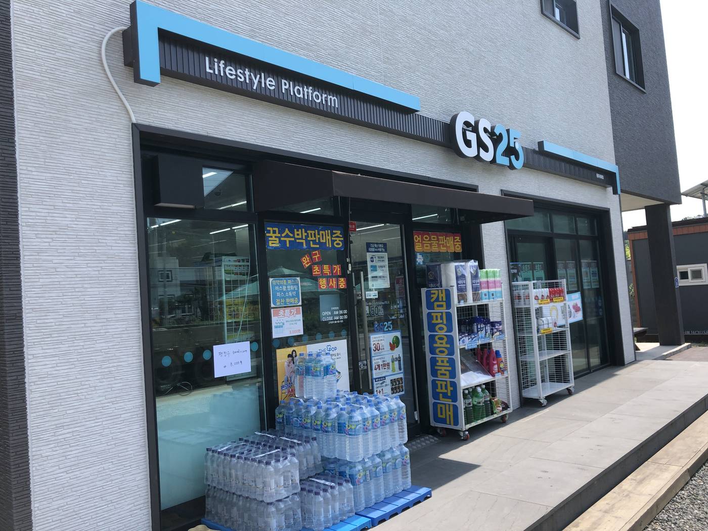 The front of a GS25 convenience store in a small Korean town: a white-and-grey building with the blue GS25 sign, stacked bottled water outside, and a door notice showing opening hours of 6 a.m. to midnight.