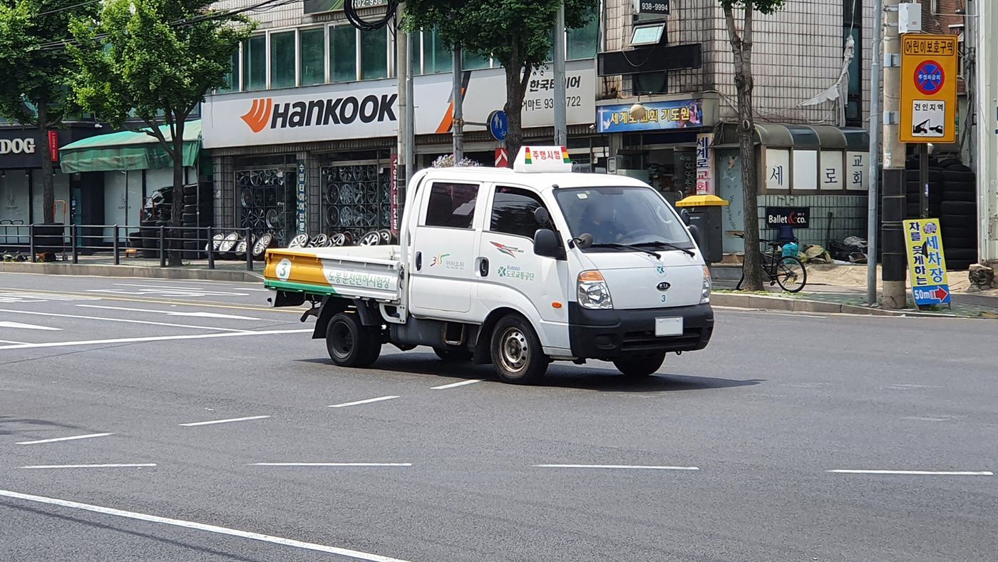 A white Kia Bongo pickup with a white 주행시험 (road test) roof sign and a yellow-and-green load bed marked 도로운전면허시험용, driving through a junction in Dobong, Seoul; the truck is used for the 1st-class ordinary licence road test.