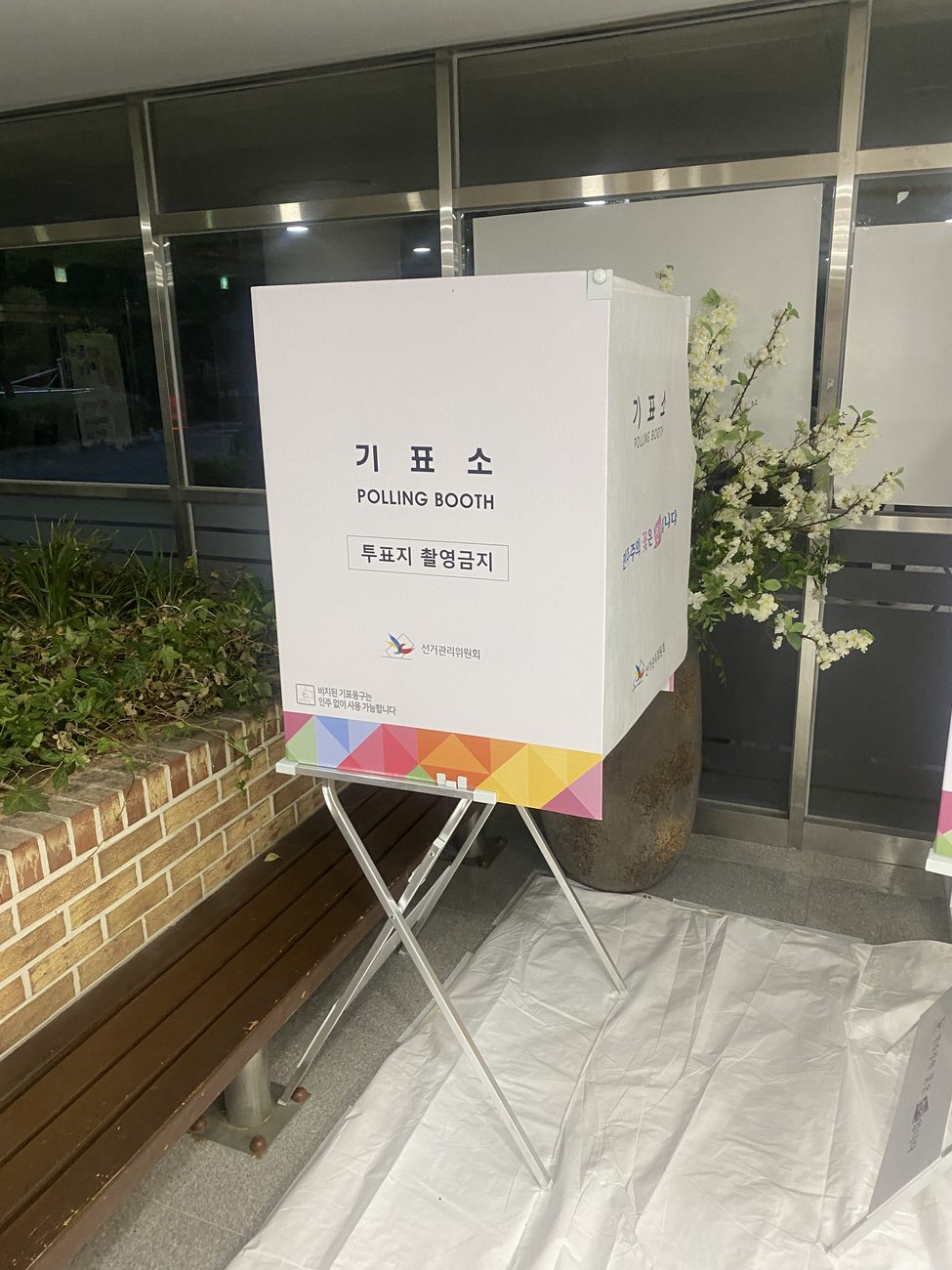 A white cardboard polling booth on a metal stand in a Korean polling station, marked 기표소 POLLING BOOTH with a sign that says photographing the ballot is prohibited (투표지 촬영금지) and the National Election Commission logo.