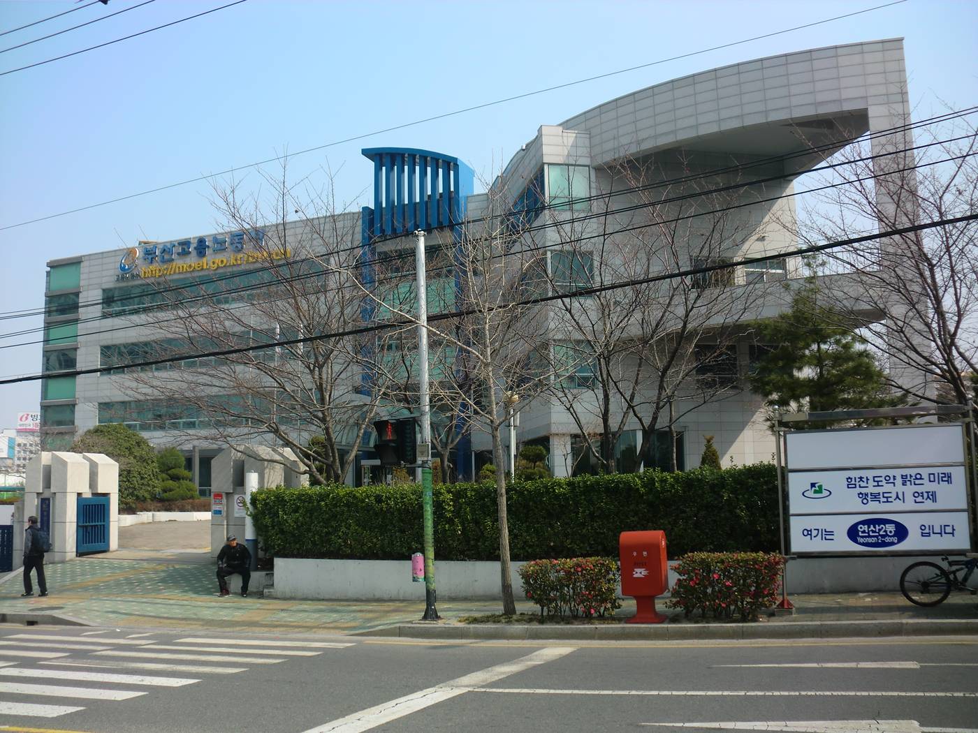 The Busan Regional Employment and Labor Office: a white and glass building with the sign 부산고용노동청 behind bare trees and a hedge, a red postbox on the pavement in front.