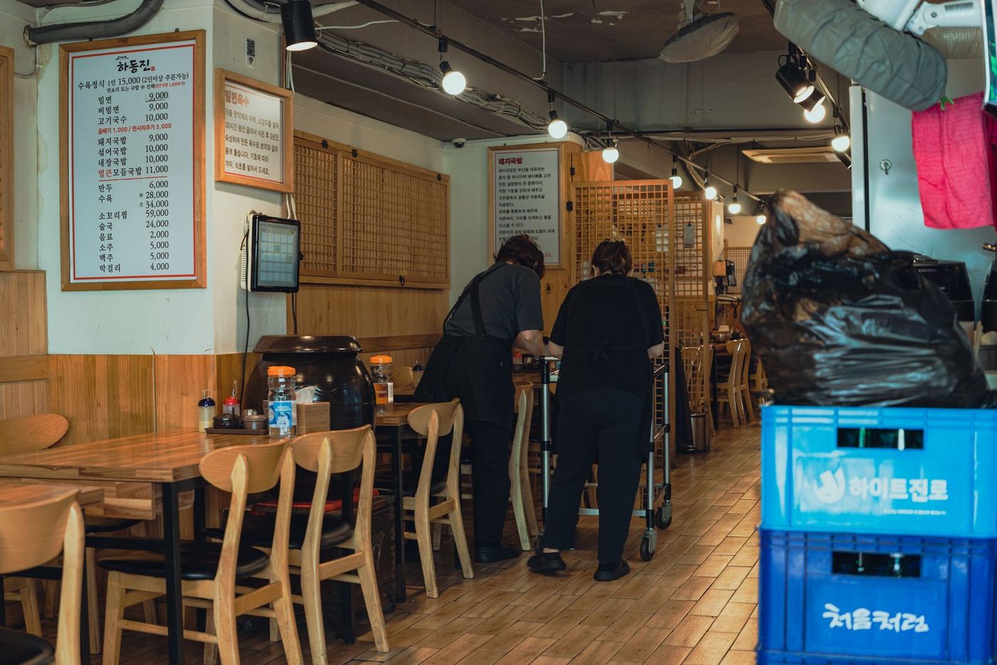 Two restaurant staff in black aprons wheel a trolley between wooden tables in a Korean restaurant; a menu board on the wall lists noodle and gukbap dishes from ₩9,000, and blue Hite Jinro and Chum Churum (처음처럼) soju crates stand in the foreground.