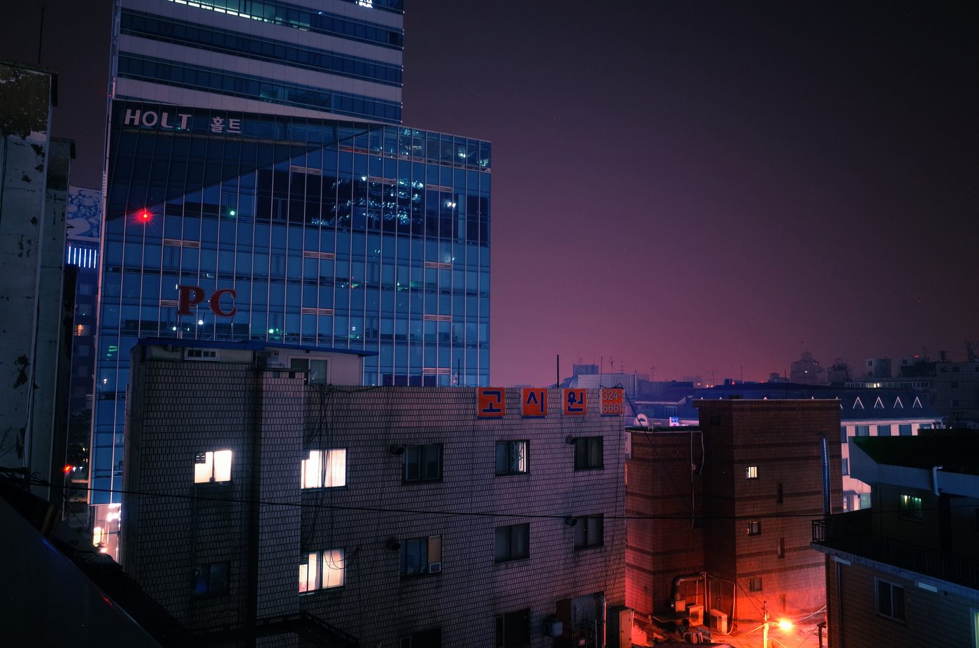 A night view of a brick residential building in Korea with lit windows and the orange sign 고시원 (goshiwon) on its roof, a phone number beside it, and a glass tower with a red PC sign behind.