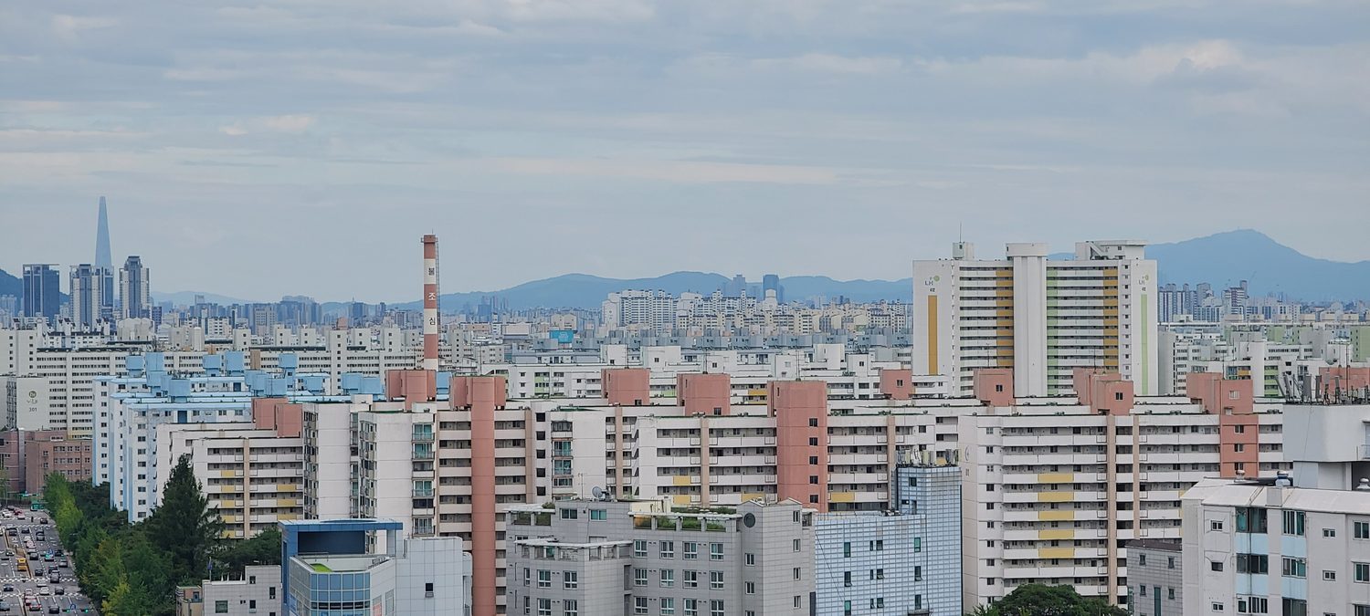A wide view of Nowon-gu, Seoul: rows of white and pink high-rise apartment blocks with numbered towers in the foreground, a red-and-white chimney, Lotte World Tower on the horizon at left and mountains behind.