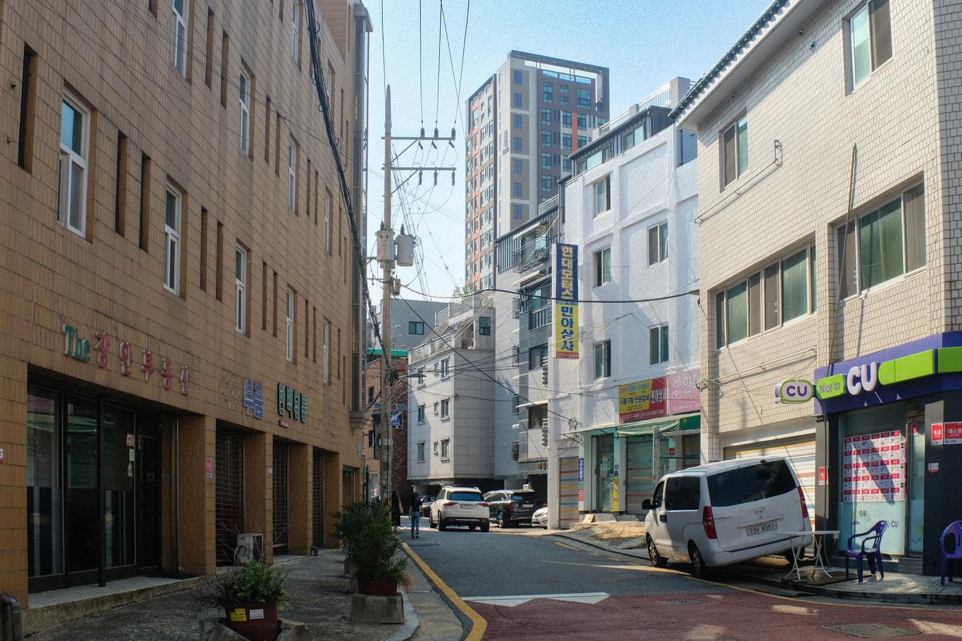A side street of four- and five-storey residential buildings in Seoul in afternoon sun: tiled and white facades, a CU convenience store with a blue-and-green sign on the right, a parked white van, overhead power lines and a newer high-rise at the end of the street.