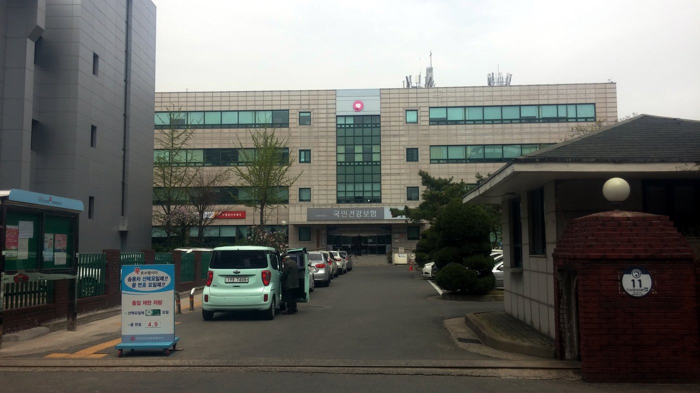 The National Health Insurance Service Bupyeong branch in Incheon: a four-storey beige office building with the NHIS logo at the top of a glass stairwell and the sign 국민건강보험 above the entrance, with cars parked in the forecourt.