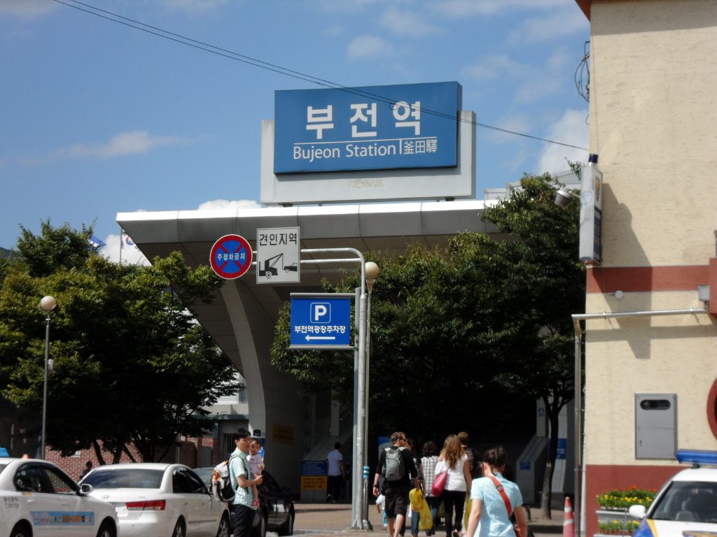 The Korail Bujeon Station building with a blue sign reading 부전역, Bujeon Station and 釜田驛 above a white canopy, with passengers walking toward the entrance, September 2010.