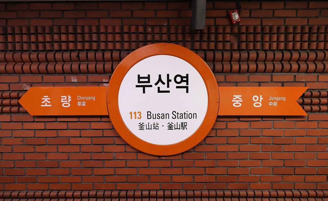 A round orange-and-white Busan Metro station sign on a brick wall reading 부산역, 113 Busan Station and 釜山站, with arrows to Choryang and Jungang.