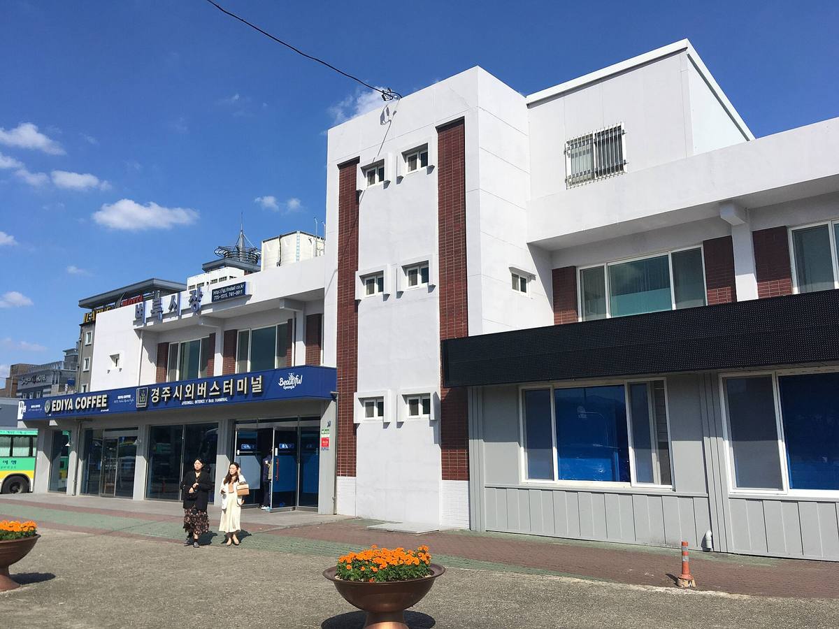 The white Gyeongju Intercity Bus Terminal with a blue sign reading 경주시외버스터미널 and Gyeongju Intercity Bus Terminal, an Ediya Coffee shop at the left end, flowerpots and two pedestrians in front under a blue sky.
