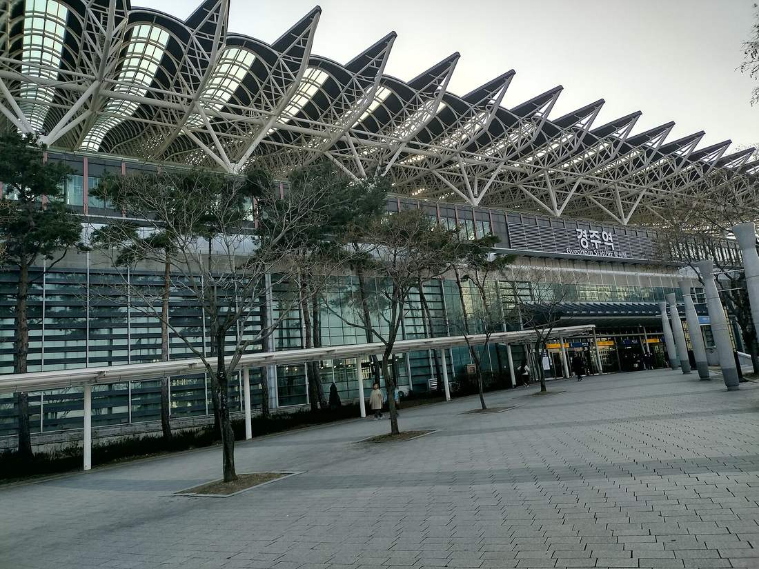 The front of Gyeongju Station, with the Korean sign 경주역 and the English Gyeongju Station under a long white steel canopy shaped like rows of roof tiles, pine trees and a paved plaza in front, March 2025.