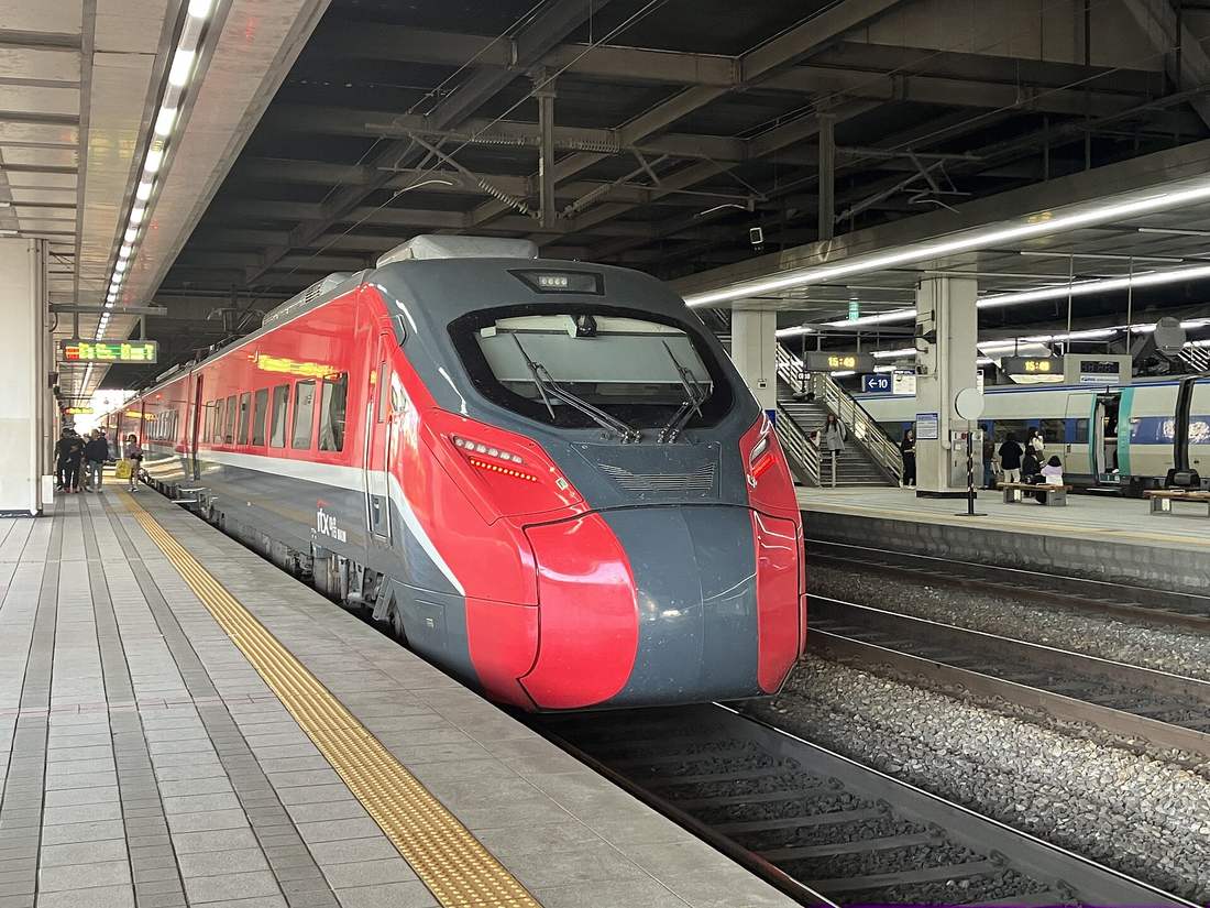 A red-and-dark-grey ITX-Maum electric train with the ITX 마음 logo beside the cab, standing at a covered platform with yellow tactile paving; another Korail train waits at the next platform.
