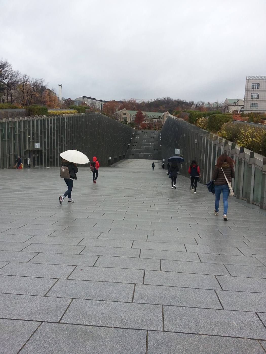 The Ewha Campus Center (ECC) at Ewha Womans University in Seoul on a rainy autumn day: a wide paved walkway sloping down between two tall glass-and-steel walls of a sunken building, students with umbrellas walking toward a stone building and autumn trees at the top of the slope.