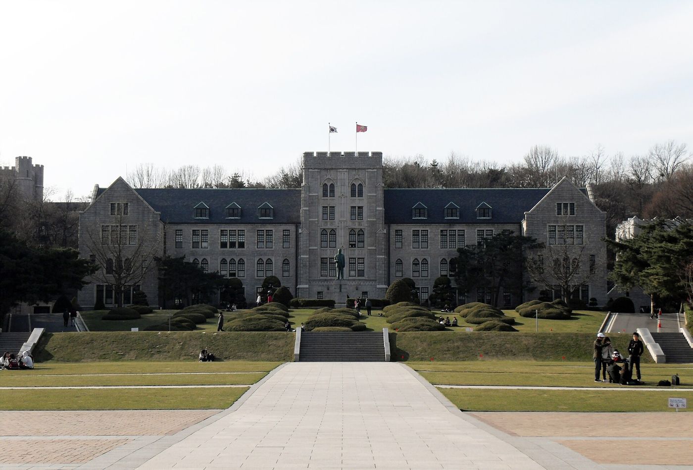Korea University's Main Hall in Seoul: a long grey granite Gothic-style building with a central tower flying two flags, a statue in front, terraced lawns and a wide stone walkway leading up to it, bare winter trees on the hill behind.