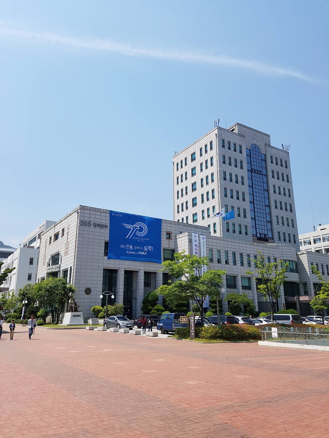 Pusan National University's main administration building in Busan: a white stone-clad block with a ten-storey tower and a blue glass stripe, a large blue banner marking the university's 70th anniversary on the lower wing, flags, trees and parked cars in front of a red-brick plaza under a blue sky.