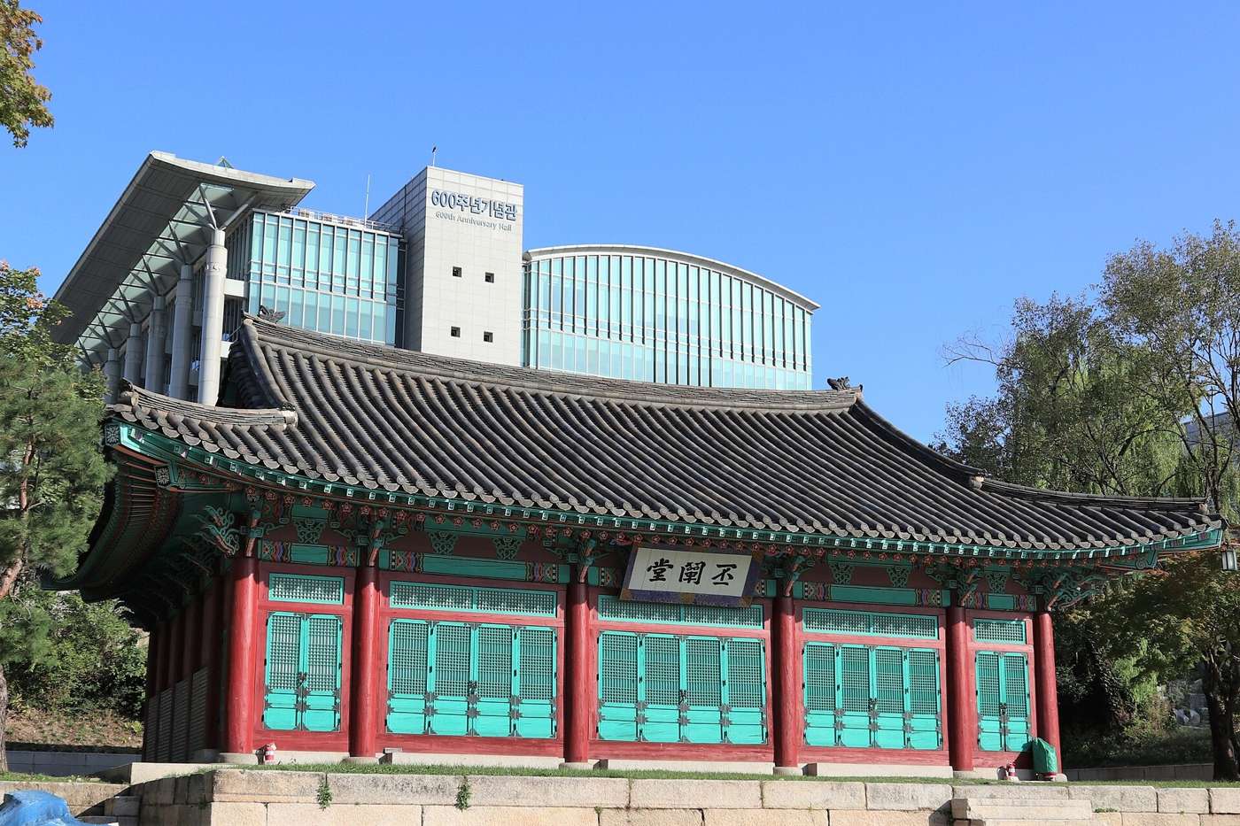 Bicheondang and the 600th Anniversary Hall at Sungkyunkwan University in Seoul: a Joseon-era wooden hall with red pillars, green latticed doors, a grey tiled roof and a signboard reading 丕闡堂, with the modern glass-and-concrete 600th Anniversary Hall rising behind it under a blue sky.