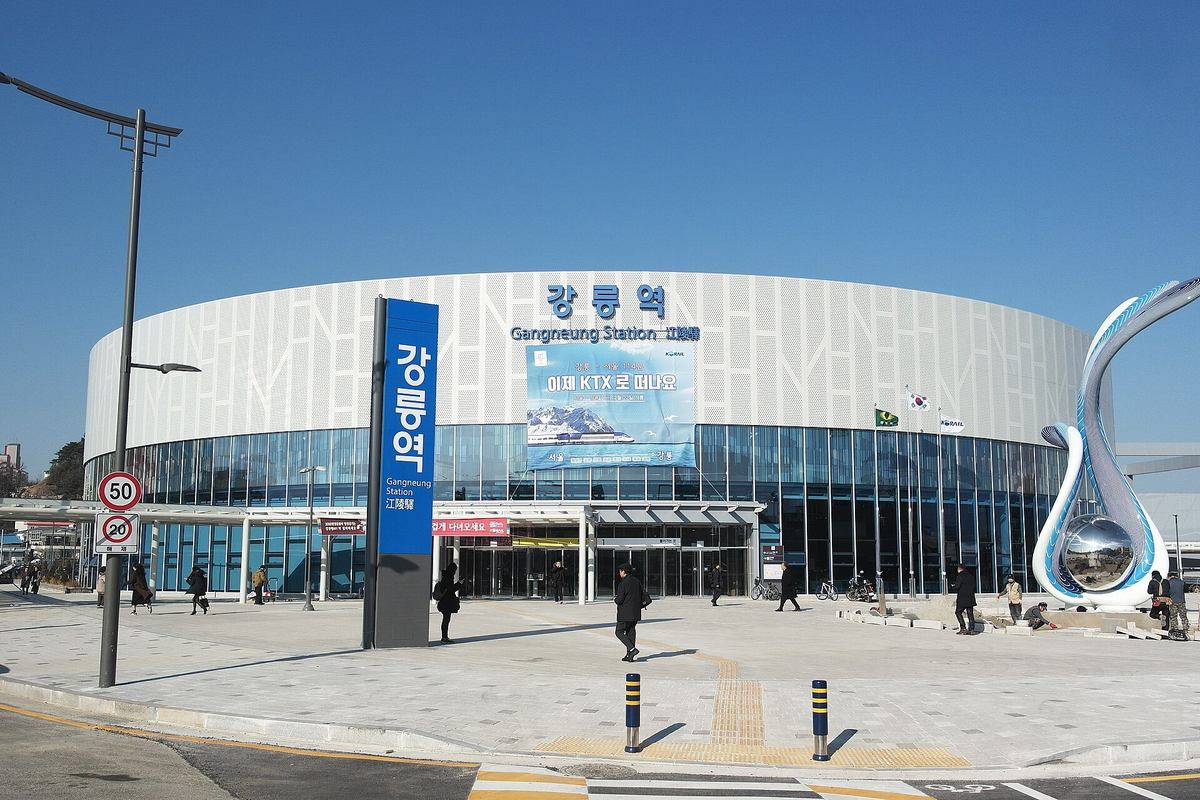 The round white Gangneung Station building with the sign 강릉역, Gangneung Station and 江陵驛, a tall blue 강릉역 pylon in front, and a Korail banner reading 강릉 – 서울 114분, 이제 KTX로 떠나요, on a clear winter day.