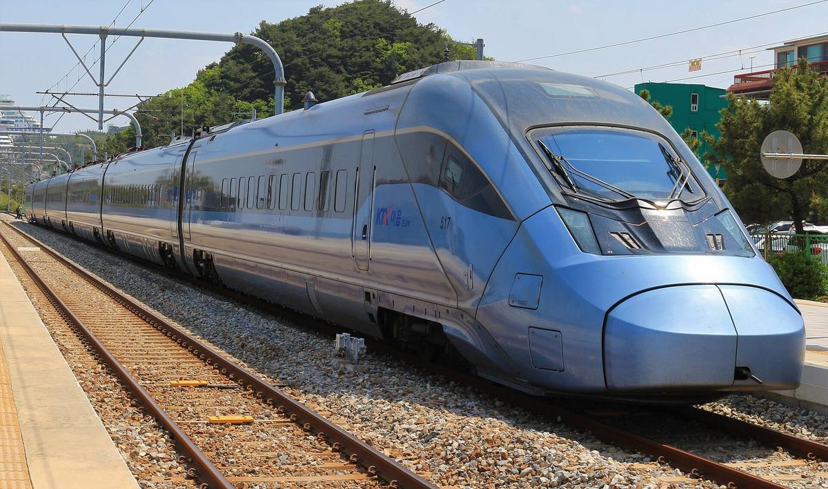 A pale blue KTX-Eum train, set 517, with the KTX 이음 EUM logo on its nose, standing at Jeongdongjin Station's platform in sunshine, pine trees on a hill behind.