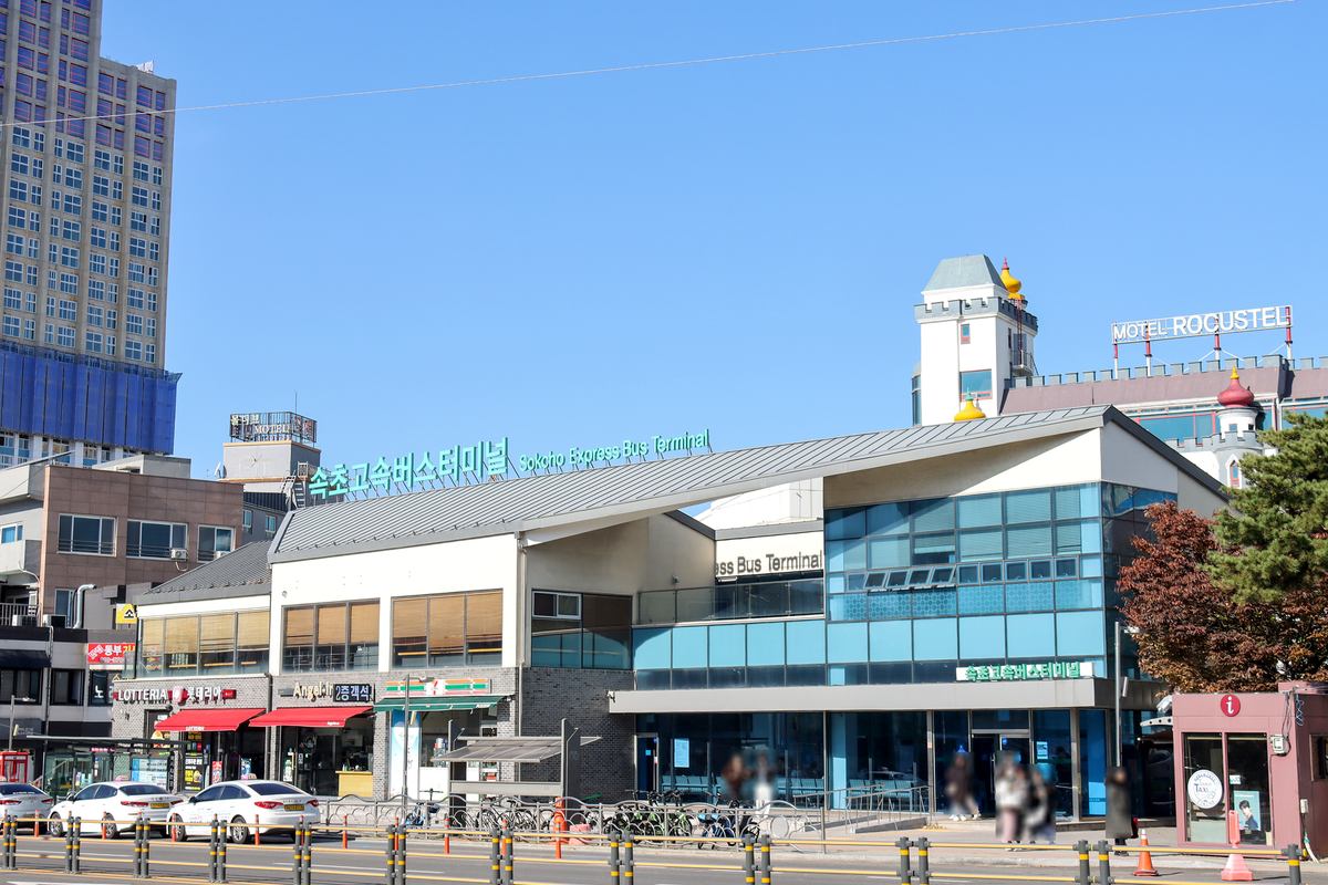 The Sokcho Express Bus Terminal, a low white-and-glass building with green lettering 속초고속버스터미널 and Sokcho Express Bus Terminal on the roof, white taxis parked along the road in front under a clear blue sky.