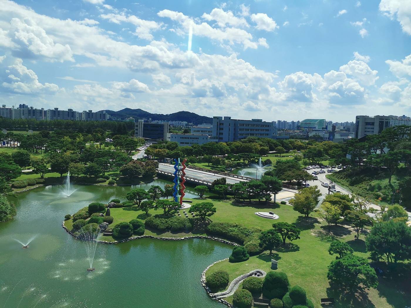 The KAIST main campus in Daejeon seen from above: a large pond with several fountains, a small island with pine trees and a red-and-blue zigzag sculpture, a bridge with a road, low campus buildings and apartment towers on the hills behind, under a blue sky with white clouds.