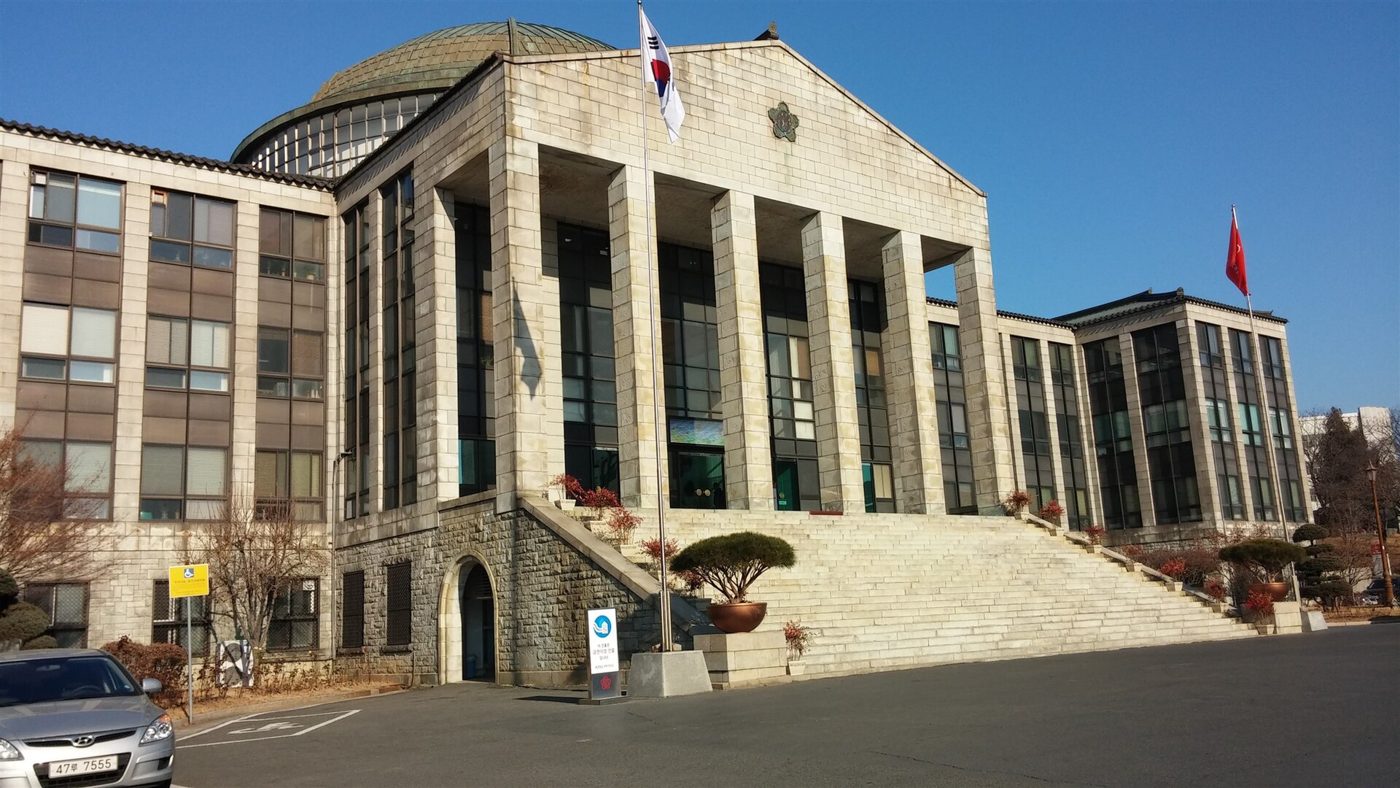 Kyungpook National University's main administration building in Daegu: a pale stone building with a tall pillared portico and a green copper dome behind, a broad flight of stone steps, the Korean flag on a pole and a red foreign flag to the right, under a clear blue winter sky.