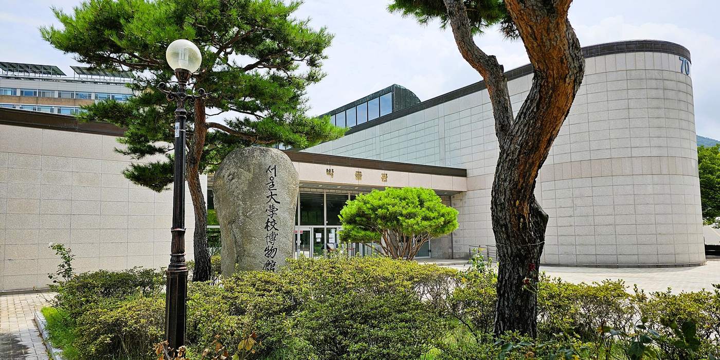 Seoul National University Museum on the Gwanak campus: a curved pale stone building with a glass roofline, a tall standing stone carved with 서울大學校博物館 in front, pine trees and a lamp post, under a bright summer sky.