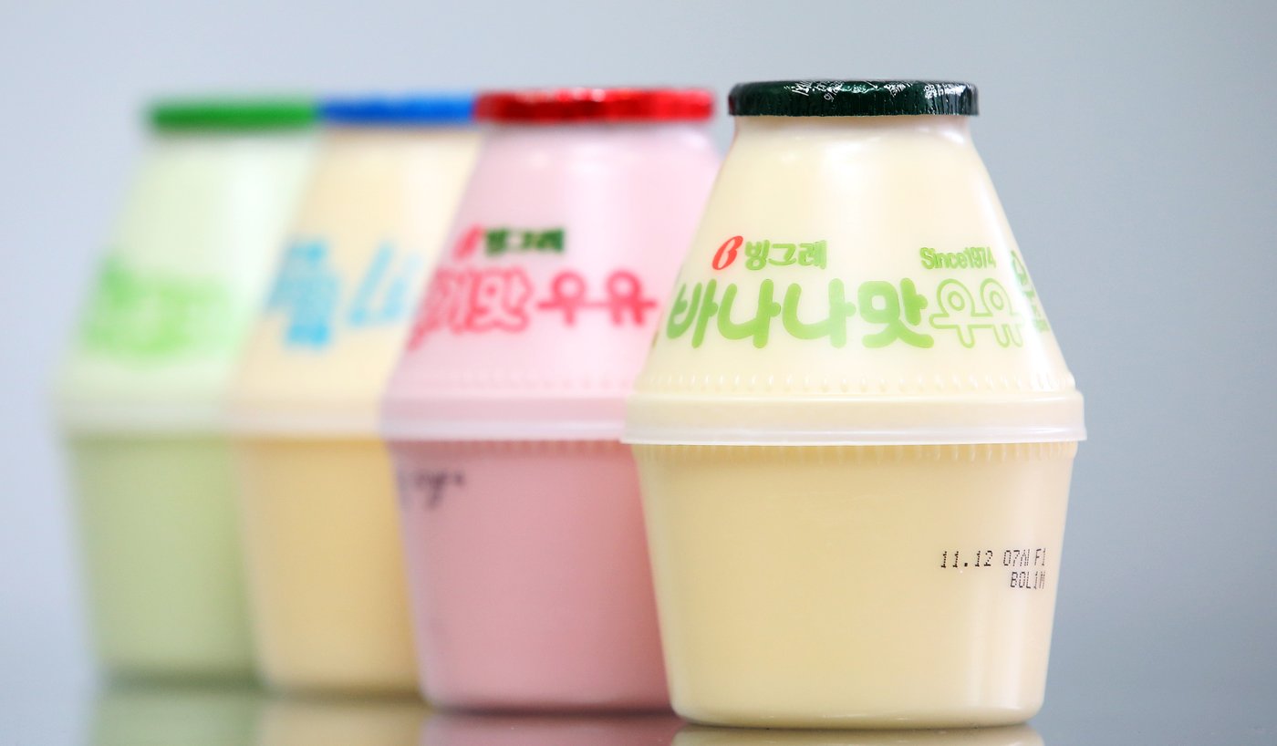 Four Binggrae flavoured milks in their squat bottles on a pale background, the banana one in front with a dark green cap and green lettering reading 빙그레 바나나맛우유 Since 1974, a pink strawberry bottle with a red cap and two more flavours blurred behind it.