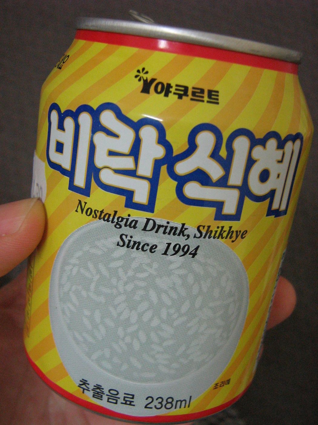 A hand holding a yellow and orange striped can of Birak Sikhye, the label reading 비락식혜 in large white letters with Nostalgia Drink, Shikhye, Since 1994 in English, a picture of rice grains, and 238ml at the bottom.