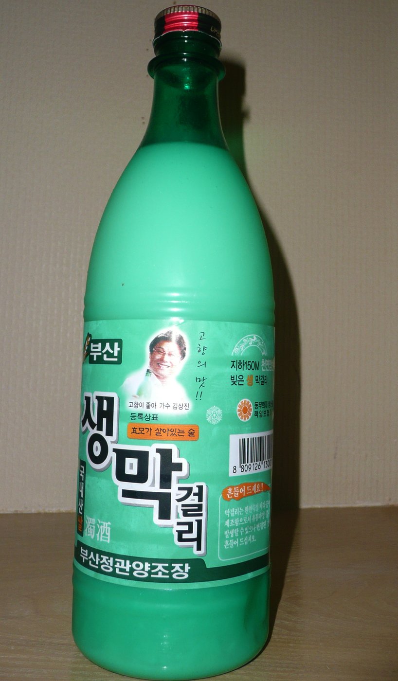 A tall green plastic bottle of Busan saeng makgeolli with a mint-green label reading 생막걸리 and 부산정관양조장, a small portrait of a singer on the label, standing against a beige wall.