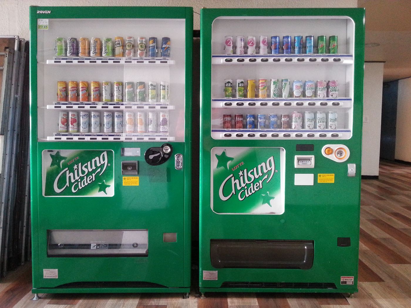 Two green Lotte Chilsung Cider vending machines side by side in a Korean building lobby, their windows lined with cans of Korean soft drinks, juices and coconut drinks, the white Chilsung Cider logo with a green star on each machine's front panel.