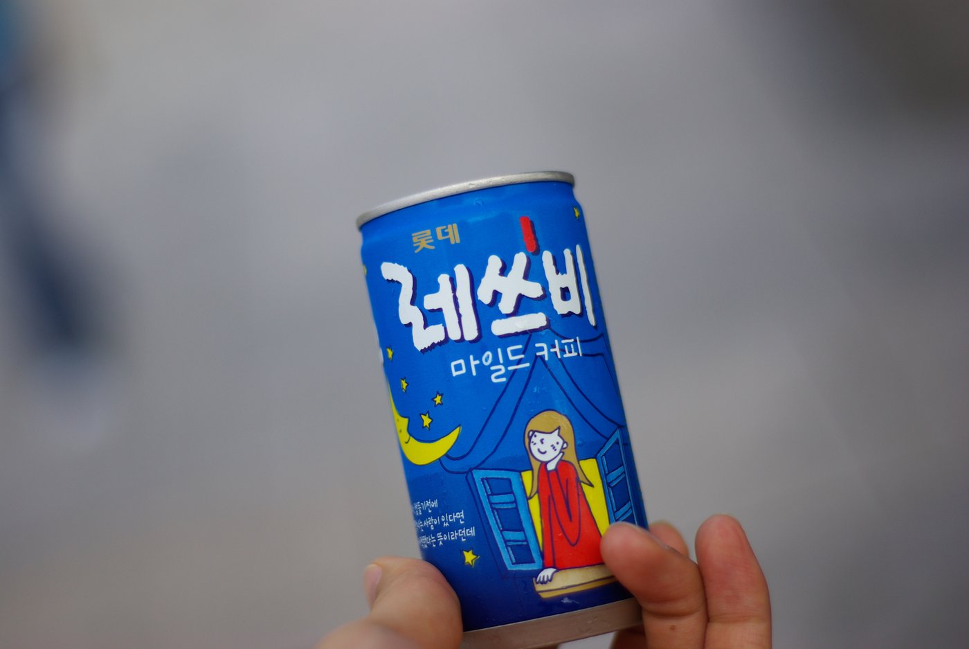 A hand holding a small blue can of Lotte Let's Be mild coffee, the label in Korean reading 롯데 레쓰비 마일드 커피 above a cartoon of a girl leaning out of a window under a crescent moon.