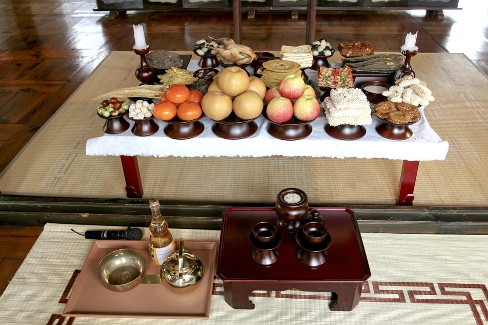 A Korean ancestral-rite table on a straw mat: wooden stands of apples, Asian pears, persimmons and tangerines, rice cakes, dried fish and a boiled chicken, white candles at both ends, and a small lacquered table with ritual cups and a brass bowl in front.