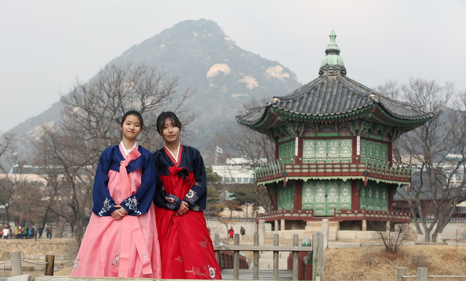 Two young women in hanbok, one pink and one red, standing on the bridge to Hyangwonjeong pavilion at Gyeongbokgung Palace in Seoul, with the green-roofed hexagonal pavilion and Bugaksan mountain behind them in winter.
