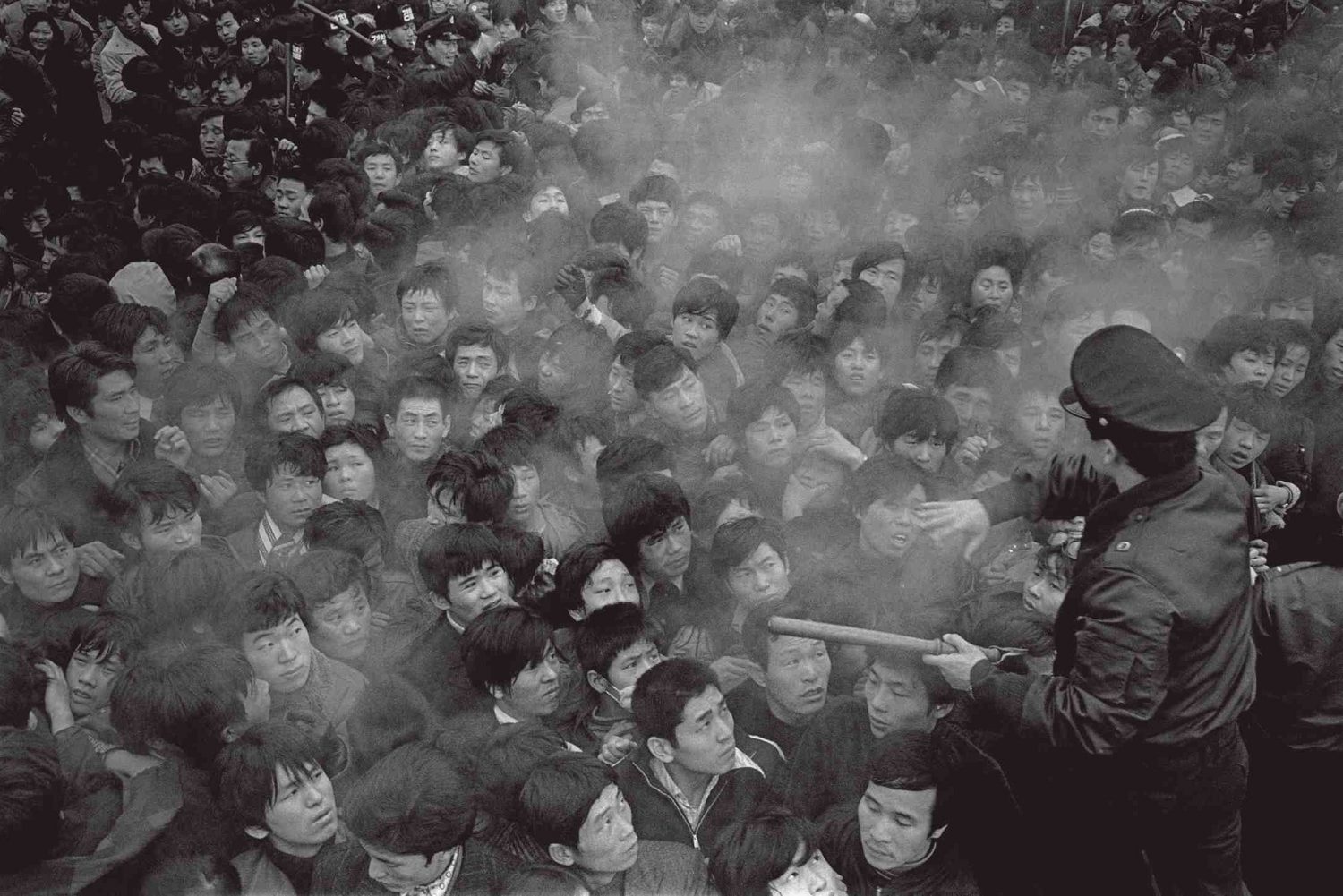 A dense crowd of young Korean men in winter coats pushing forward at a Seoul station in 1978 while a uniformed station attendant holds them back with a baton; a haze hangs over their heads.
