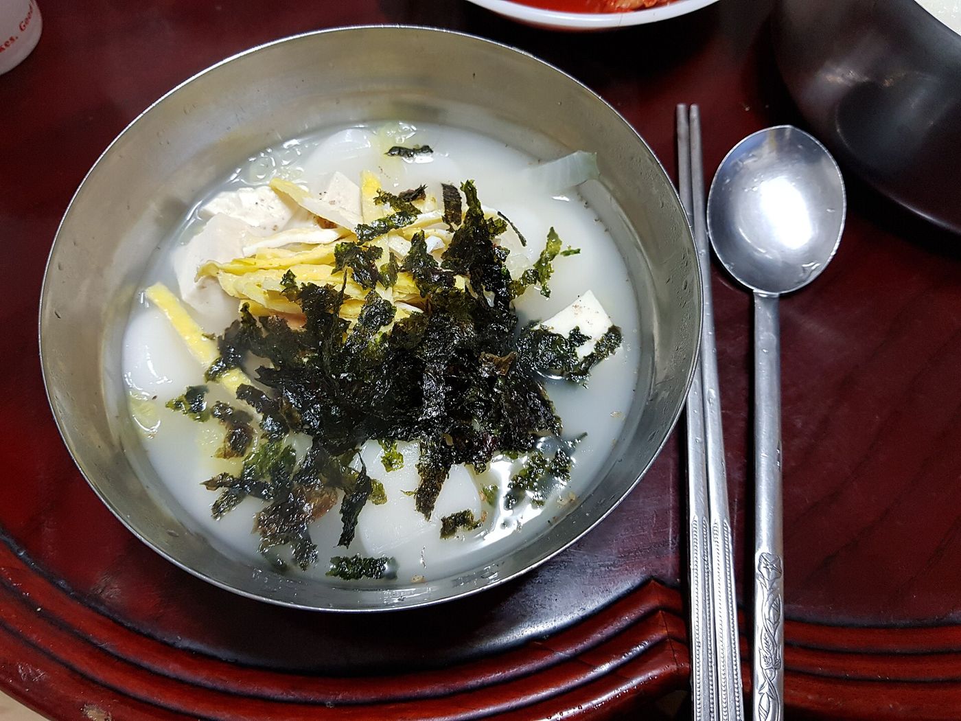 A stainless-steel bowl of tteokguk, sliced rice-cake soup in a milky broth topped with shredded egg and crumbled seaweed, with a spoon and chopsticks on a dark wooden tray.