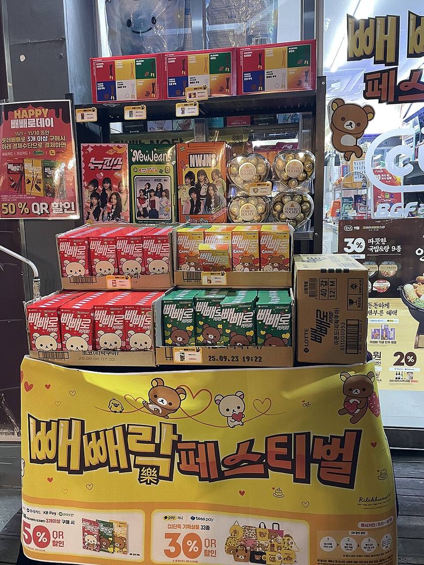 A CU convenience store's Pepero Day display in Korea, November 2024: Rilakkuma-themed Pepero boxes priced ₩1,800, NewJeans Pepero sets, Ferrero Rocher hearts, a six-flavour Pepero box on the top shelf, and signs advertising 50% QR discounts with Korean cards and pay apps.