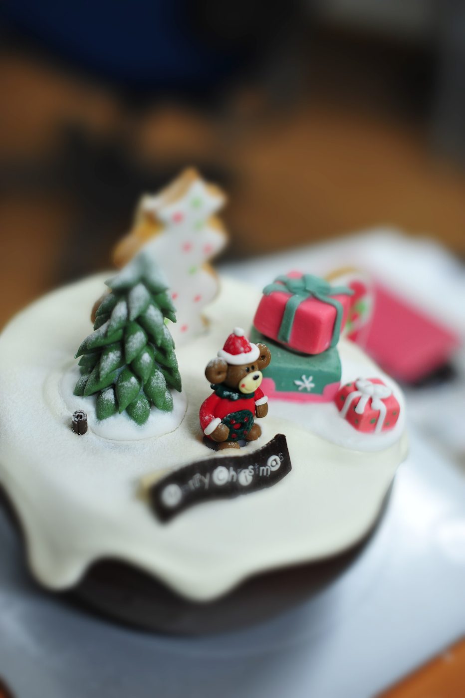 A small Korean Christmas cake with white icing drizzled over chocolate sponge, topped with a sugar-paste fir tree, a bear in a Santa hat holding a wreath, wrapped gifts and a chocolate plaque reading Merry Christmas.