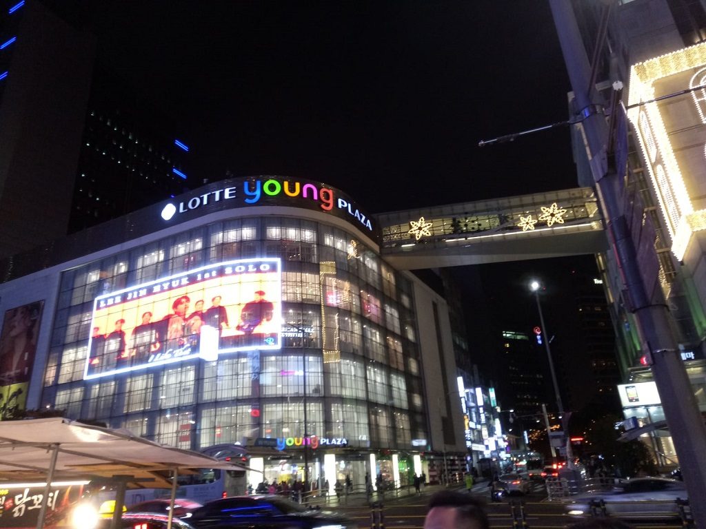 The curved glass façade of Lotte Young Plaza in Myeongdong at night with its rainbow "young" sign, a lit advertising screen on the front, and gold star lights on the pedestrian bridge beside it.