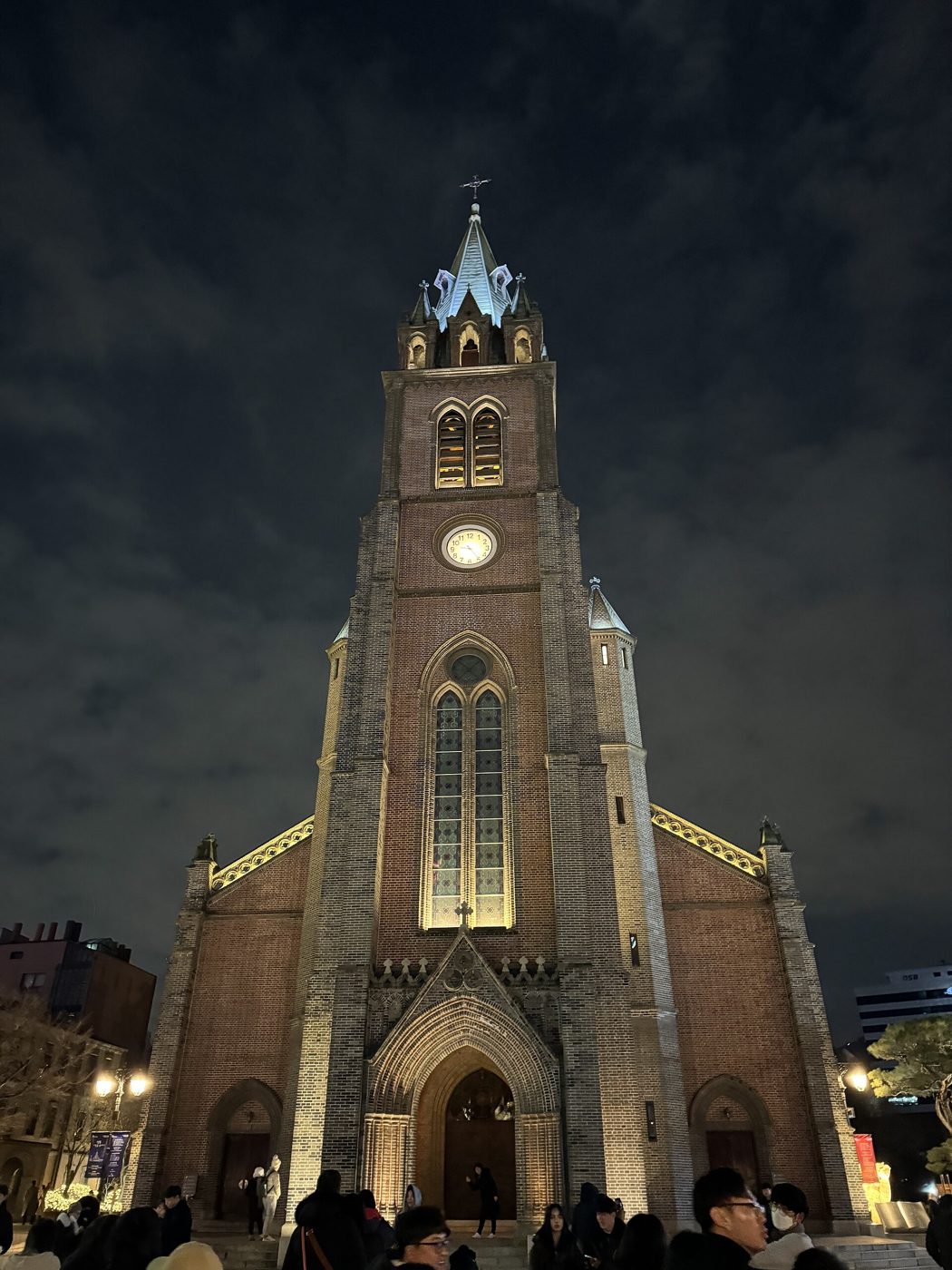 Myeongdong Cathedral at night in December, its red-brick Gothic tower and spire floodlit, a crowd in winter coats gathered in front of the main door.