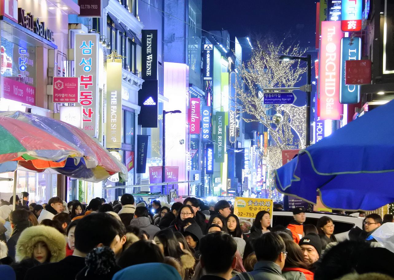 A dense crowd in winter coats filling Myeongdong's main street at night, neon shop signs in Korean and English on both sides, a tree wrapped in white Christmas lights over the street.