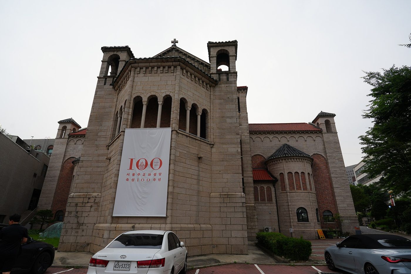 The stone and red-brick Seoul Anglican Cathedral in Jeong-dong with its arcaded bell towers and a large white banner marking 100 years since its consecration, 1926 to 2026.