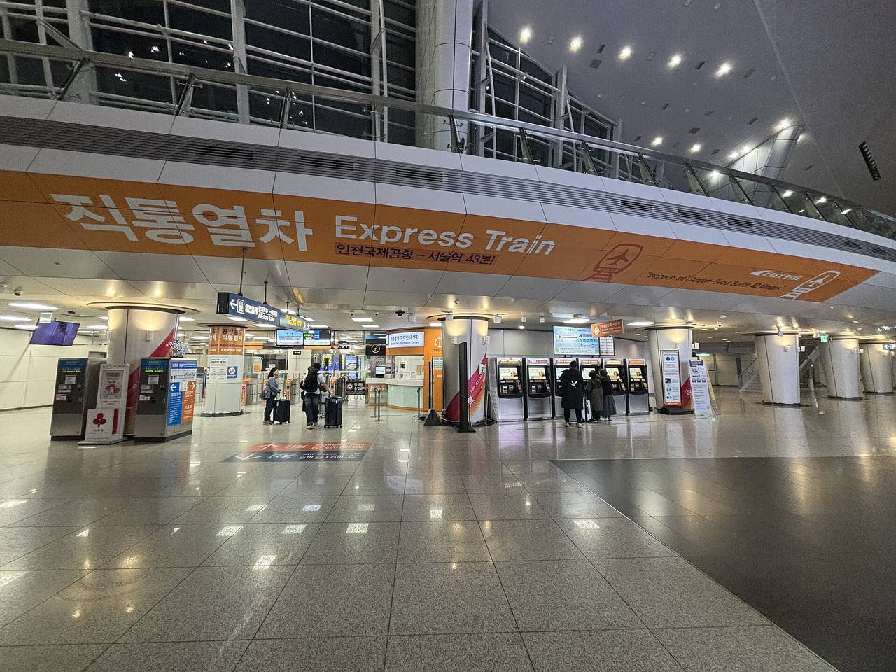 The Airport Railroad express train concourse at Incheon Airport Terminal 1 station: an orange overhead band reading 직통열차 Express Train and 인천국제공항–서울역 43분, with ticket machines, an information desk and travellers with suitcases at the gates.