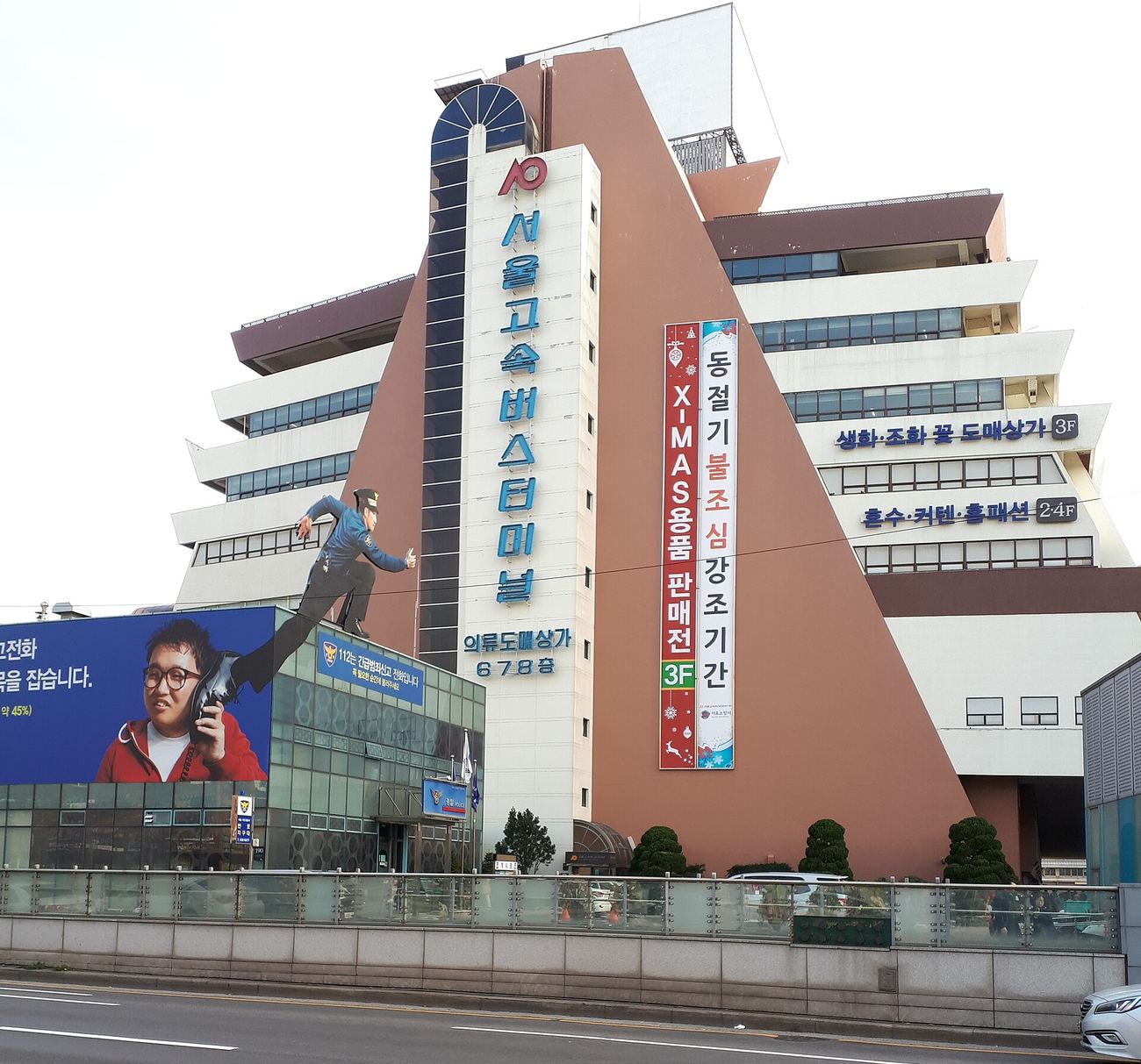 The Seoul Express Bus Terminal building in Seocho-gu, its tall white tower carrying the Hangul sign 서울고속버스터미널, with the stepped brown-and-white terminal block behind and a police box and road in front.