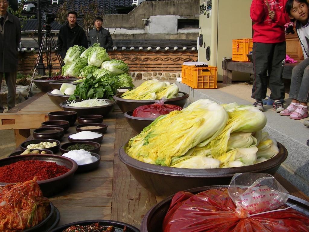 A wooden table in a hanok courtyard laid with bowls of salted cabbage, chilli flakes, garlic, salt and other kimchi ingredients, with children watching.